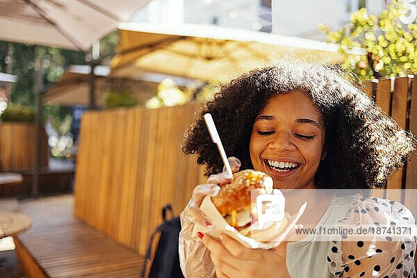 Junges afrikanisches amerikanisches Mädchen ißt klassischen Burger und trinkt Limonade in der Stadt. Outdoor Frau trägt Sommerbluse mit Tupfen eatting leckeren Burger beim Sitzen auf der Terrasse. Frau in blau Verkostung Sandwich mit freudigen Blick bewundern sonnigen w