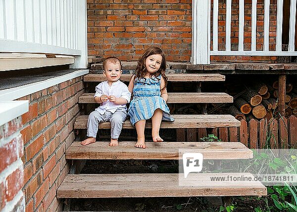 Niedliche kleine Bruder und Schwester sitzen auf Holztreppe in der Nähe Veranda des Landhauses und entspannen in der ländlichen Gegend während der Sommerferien