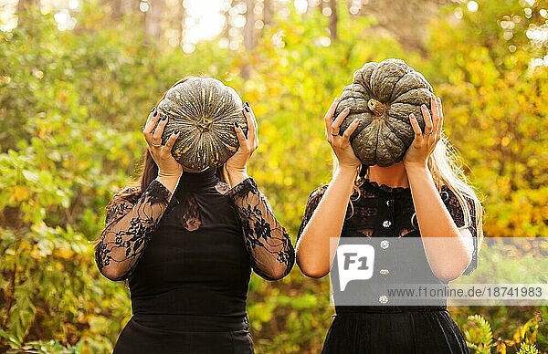 Zwei Freundinnen  die gruselige grüne Kürbisse vor ihrem Gesicht halten  stehen isoliert über Herbstwald Hintergrund. Herbstferien  halloween Dekorationen und Menschen Konzept