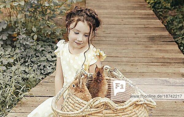 Kleines hübsches Mädchen im Sommerkleid  das draußen auf einem Holzweg vor einem Strohkorb mit Kaninchen sitzt  süßes Kind  das die Kaninchen mit einem leichten Lächeln betrachtet und Zeit mit ihren Lieblingstieren genießt