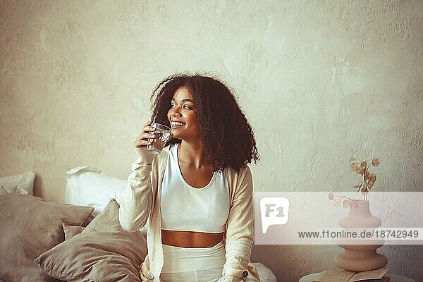 Aqua Balance. Junge lächelnde afroamerikanische Frau sitzt zu Hause mit einem Glas sauberem Mineralwasser  glückliches lockiges schwarzes Mädchen genießt gesunde Morgenroutine. Aufrechterhaltung des Flüssigkeitshaushalts