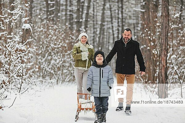 Fröhliche Eltern  die hinter einem fröhlichen Jungen mit Schlitten gehen  während sie an einem Wintertag Zeit im verschneiten Wald verbringen