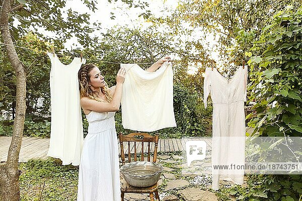 Junge Frau hängt Wäsche im Freien auf. Nettes Mädchen im Kleid wäscht weiße Wäsche in einem Metallbecken im Hinterhof  hängt Wäsche auf Wäscheleine und lässt sie im Garten trocknen  trocknet Kleidung im Freien im Wind