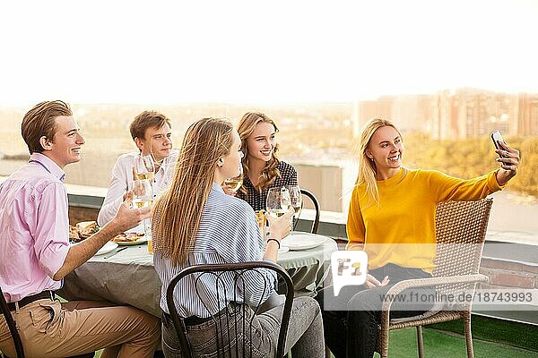 Gruppe von fünf fröhlichen jungen Menschen beim Abendessen im Restaurant  sitzen auf der Terrasse am Tisch und halten Gläser Wein in den Händen posieren für selfie auf Smartphone. Freundschaft und Versammlung Konzept