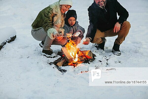 Fröhliche Eltern und Sohn genießen ein heißes Getränk  während sie sich abends am Lagerfeuer im Winterwald ausruhen