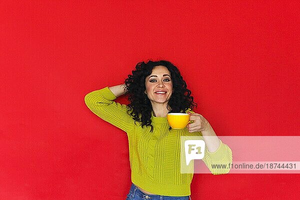Junge glückliche lockige Frau mit einer Tasse Tee in der Hand  die isoliert an einer roten Wand posiert. Lächelnde zufriedene Frau  die ein heißes Getränk genießt und Kaffee aus einem Becher trinkt  während sie vor einem hellen Studiohintergrund steht