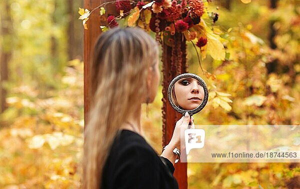Scenic Foto von jungen schönen Mädchen mit Abendmakeup trägt schwarze Hexe Kostüm hält Spiegel und Blick auf sie während Halloween Foto Session im Herbst Wald. Saisonale Feiertage Konzept