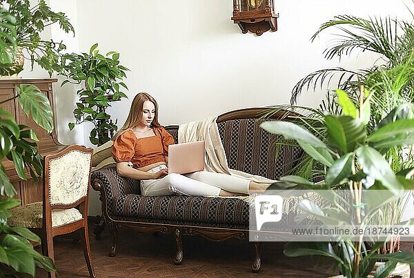 Barfüßige Frau benutzt ein Netbook und entspannt sich auf einer eleganten Couch inmitten üppiger Topfpflanzen