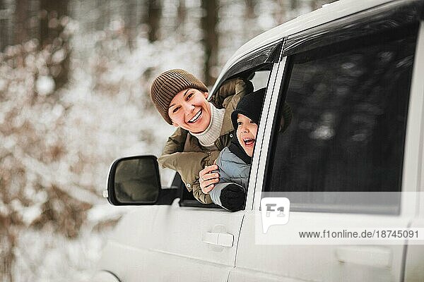 Fröhliche erwachsene Frau und kleiner Junge in Oberbekleidung  die in die Kamera schauen  während sie aus dem Fahrzeugfenster schauen  vor einem unscharfen Hintergrund eines Winterwaldes
