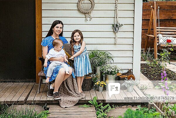 Junge begeisterte Mutter sitzt im Sessel mit entzückenden kleinen Geschwistern auf der Terrasse eines Landhauses und entspannt sich gemeinsam am Wochenende