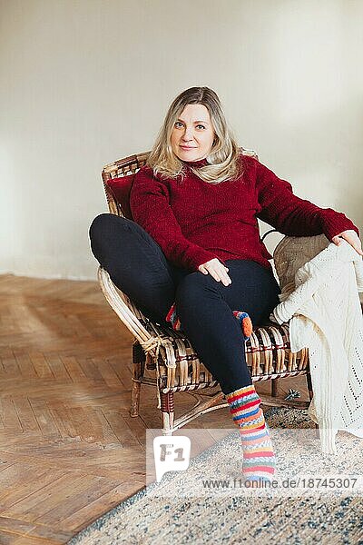 Attraktive glückliche Frau mittleren Alters  die am Wochenende zu Hause auf einem bequemen Stuhl sitzt und sich entspannt. Sie trägt bunte Stricksocken und einen warmen roten Pullover und lächelt in die Kamera. Menschen Lebensstil Lebensstil