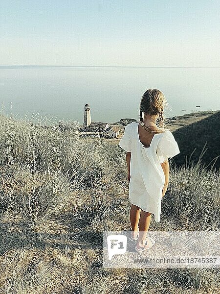 Ganzkörper Rückenansicht eines einsamen  nicht erkennbaren Mädchens im weißen Sommerkleid  das auf einem grasbewachsenen Hügel an der Meeresküste mit einem Leuchtturm steht