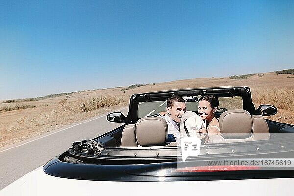 Rückansicht eines glücklichen Paares mit Hüten  das die Freiheit genießt  während es in einem Cabrio durch eine grasbewachsene Wüstenlandschaft im Sommer fährt