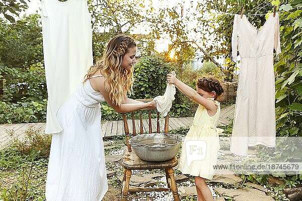 Charmantes kleines Mädchen  das seiner Mutter mit einem Weidenkorb hilft  während es im Sommerabend im Garten die Wäsche aufhängt