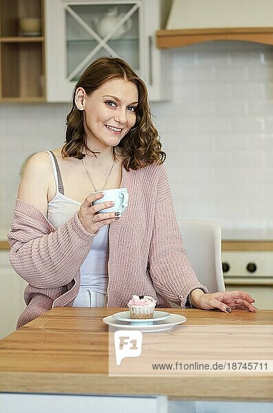 Glückliche junge Frau ißt leckeren rosa Muffin mit einem breiten Lächeln und geschlossenen Augen  nachdem sie einen Bissen genommen und probiert hat  und hält eine Tasse mit einem heißen Getränk über dem Küchenhintergrund. Ungesunde Lebensmittel Konzept