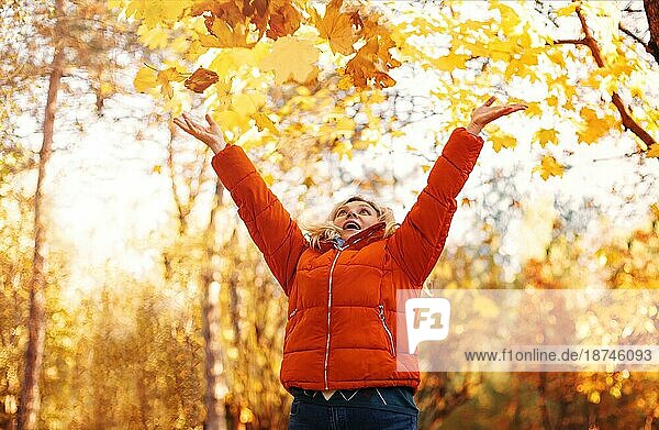 Zufriedene Frau in Oberbekleidung lächelnd mit trockenen Blättern an einem sonnigen Wochenendtag im Herbstpark
