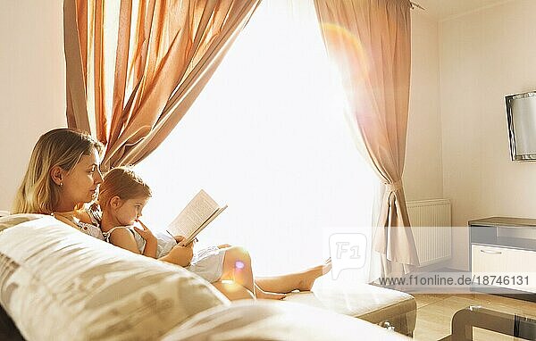 Entspannte Mutter und kleine Tochter sitzen zusammen auf einem gemütlichen Sofa im Wohnzimmer mit einem Buch in der Hand. Die Mutter und das süße Mädchen verbringen ihre Zeit sinnvoll mit Lesen und Lernen und genießen das Wochenende zu Hause
