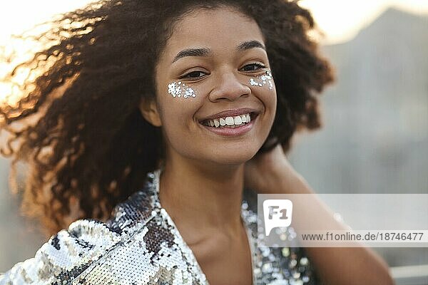 Close up Porträt von überglücklich charmante afrikanische amerikanische Frau mit lockigem Haar und Glitter auf den Wangenknochen lächelnd glücklich in die Kamera während der Bewegung tanzen im Freien  festliche Atmosphäre in der Luft
