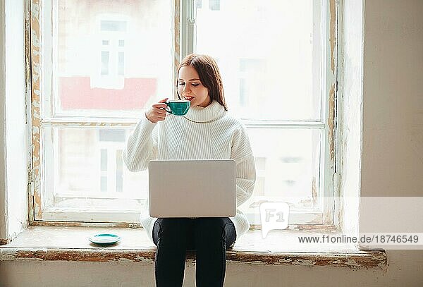 Fröhliche junge Frau genießt frischen Kaffee und surft auf ihrem Netbook  während sie auf der schäbigen Fensterbank vor dem Fenster sitzt