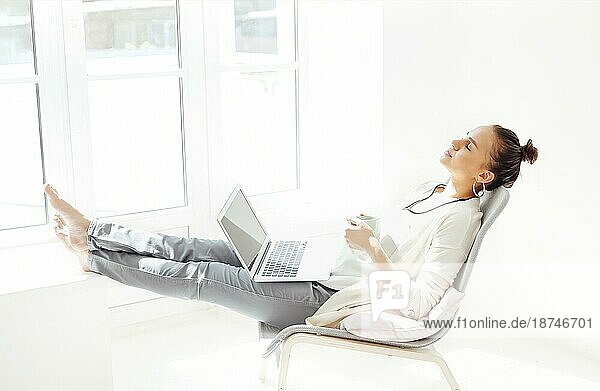 Junge Frau mit Händen hinter dem Kopf auf einem Stuhl sitzend und mit geschlossenen Augen entspannend  nachdem sie einen Laptop in einem hellen Raum zu Hause benutzt hat