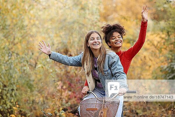 Zwei junge glückliche verschiedene weibliche beste Freunde fahren Fahrrad zusammen im Herbst Wald  aktive positive multirassische weibliche Freunde lächelnd in die Kamera und wavinh Hände beim Fahren Stadt Fahrrad im Herbst Natur