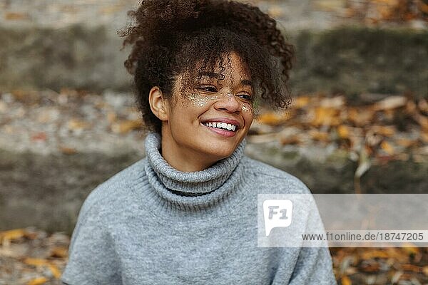 Porträt einer jungen  schönen  glücklichen afroamerikanischen Frau mit glamourösem  glänzendem Make up und in einem warmen Pullover  die bei bewölktem Herbstwetter im Freien sitzt und breit und fröhlich lächelnd in die Ferne schaut