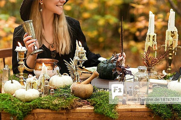 Junge Frau im Hexenkostüm sitzt am Tisch mit magischen Zutaten an einem Herbsttag im Wald. Nahaufnahme