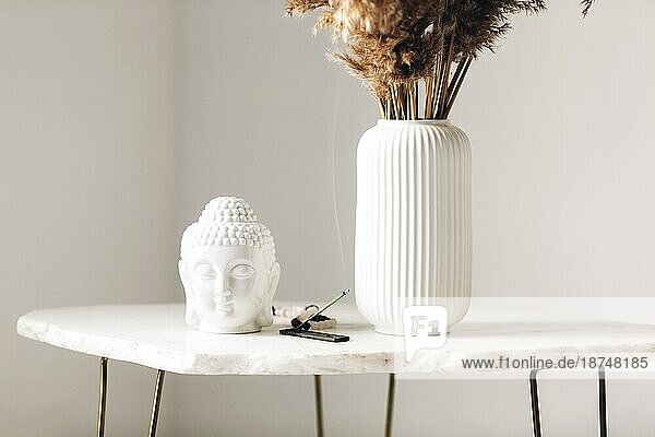 Kleiner Marmortisch mit Dekorationen  weiße Vase mit Trockenblumen  kleine Buddha Kopf Statue und brennendes Räucherstäbchen auf Ständer in modernem  einfachem Wohnungsumfeld