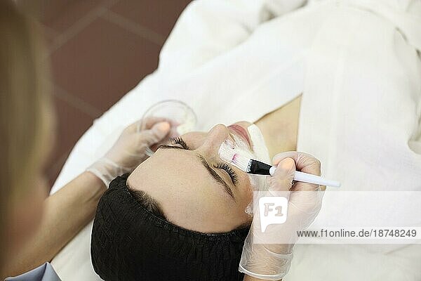 Eine nicht erkennbare Kosmetikerin trägt eine Tonmaske mit einem Pinsel auf das Gesicht einer entspannten Kundin auf  während sie eine Hautpflegeprozedur in einem SpaSalon durchführt