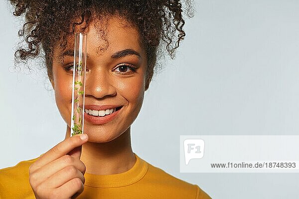 Lächelnde afroamerikanische Frau  die ein Glasreagenzglas mit Microgreens vor dem Gesicht hält  während sie vorgrauem Hintergrund posiert  Platz für Text. Superfood und Ernährung Konzept