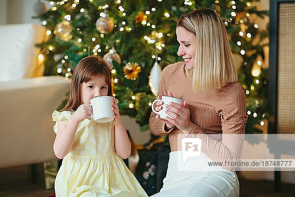 Glückliche Familie  Mutter und kleine Tochter trinken heißen Kakao  sitzen neben dem geschmückten Weihnachtsbaum  Mutter und Kind verbringen Zeit zusammen zu Hause während der Winterferien und feiern das neue Jahr