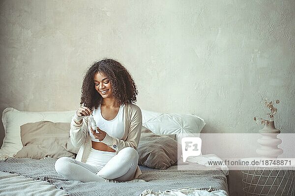 Schönheit und Hautpflege Konzept. Cropped Foto von afrikanischen amerikanischen jungen weiblichen Modell hält Flasche mit feuchtigkeitsspendende Essenz Serum sitzt gegen unscharfen modernen Wohnung Innenhintergrund