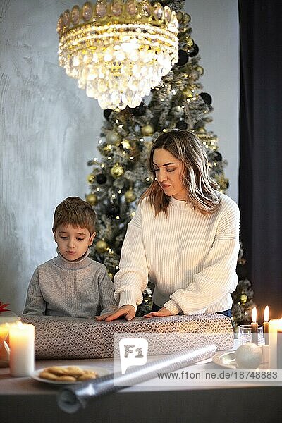 Mutter und Sohn verpacken gemeinsam Weihnachtsgeschenke zu Hause  während sie hinter einem Tisch in der Nähe eines geschmückten Tannenbaums stehen  ein kleines Kind hilft seiner Mutter beim Einpacken der Geschenke. Familientraditionen während der Winterferien