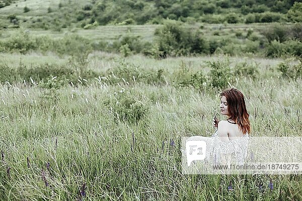 Seitenansicht einer jungen Frau  die ihr rothaariges Haar berührt und sich ausruht  während sie im Gras an einem Sommertag in der Natur sitzt
