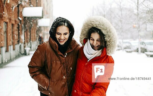 Zwei glückliche Freundinnen genießen das verschneite Wetter im Freien  Frauen beste Freundinnen in Winterkleidung Daunenjacken lachen und haben Spaß beim Spaziergang auf Stadtstraße im Winter während der Weihnachtsferien