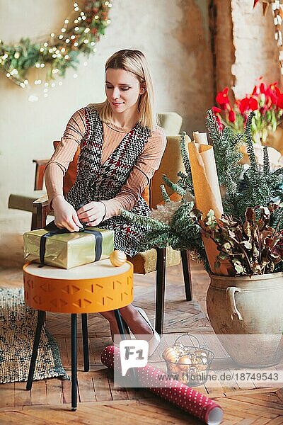 Lächelnde glückliche blonde Frau sitzt auf einem Stuhl im neuen Jahr festlich dekorierten Wohnzimmer und Verpackung Weihnachtsgeschenkbox mit Geschenkpapier und Band Dekor. Konzept der Vorbereitung für die Feiertage