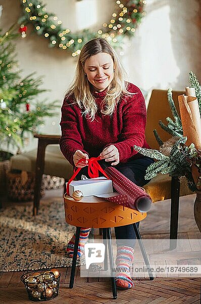 Lächelnde glückliche blonde Frau mittleren Alters  die auf einem Stuhl in einem festlich geschmückten Wohnzimmer sitzt und eine Weihnachtsgeschenkbox mit Geschenkpapier und Banddekor verpackt. Konzept der Vorbereitung für die Feiertage
