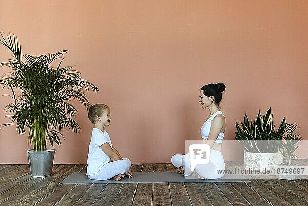 Seitenansicht einer lächelnden Frau und eines kleinen Mädchens  die eine Yogapose einnehmen  während sie auf einer Matte im gemütlichen Wohnzimmer stehen und wegschauen