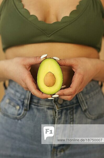 Junge Frau  die in die Kamera schaut und die Hälften einer reifen Avocado vor einem beigen Hintergrund zeigt