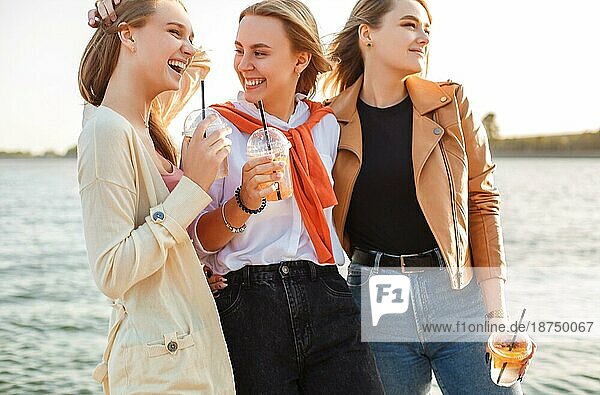 Glückliche junge Frauen mit Getränken zum Mitnehmen  die auf dem Bürgersteig spazieren gehen und an einem sonnigen Wochenendtag in der Stadt lächeln