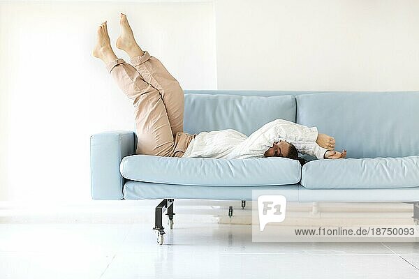 Foto einer jungen  fröhlichen Frau in Freizeitkleidung  die sich mit hochgelegten Beinen auf ein blaues  gemütliches Sofa fallen lässt und spielerisch unter ihrem Arm in die Kamera blickt  um die freie Zeit zu Hause zu genießen und Spaß mit niemandem zu haben