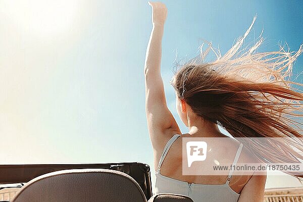 Rückenansicht einer fröhlichen jungen Frau in Sommerkleidung  die auf dem Beifahrersitz eines Cabriolets sitzt und während einer Autofahrt über die Schulter in die Kamera schaut