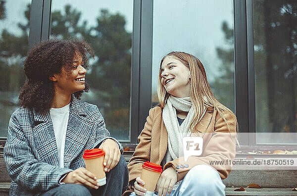 Zwei glückliche multirassische Teenagermädchen mit Kaffee zum Mitnehmen sitzen im Freien an einem Herbsttag  reden und lachen. Lächelnd multiethnischen besten weiblichen Freunde verbringen Zeit zusammen an der frischen Luft im Herbst Zeit