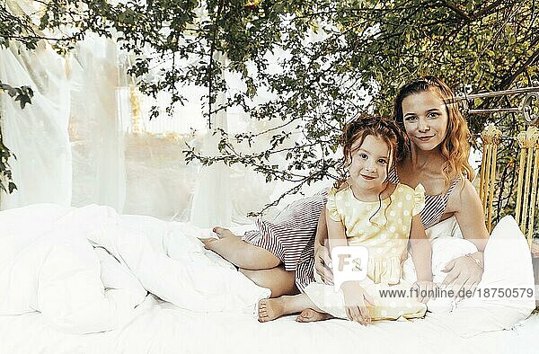 Junge schöne glückliche Familie  lächelnde Mutter und kleines Mädchen Tochter sitzt auf dem Bett mit sauberen weißen Bettwäsche in grünen Garten im Freien  Mutter und Kind genießen Sommertage  Entspannung in der Natur