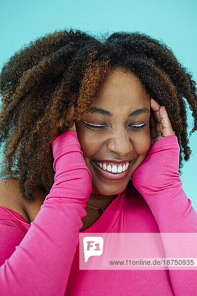 Verträumte junge schöne afroamerikanische Frau mit hellen Augenlinien  die einen rosafarbenen Body über einer blaün Wand trägt  hält die Hände unter dem Kinn zusammen  schaut mit glücklichem Gesichtsausdruck  hat ein zahniges Lächeln