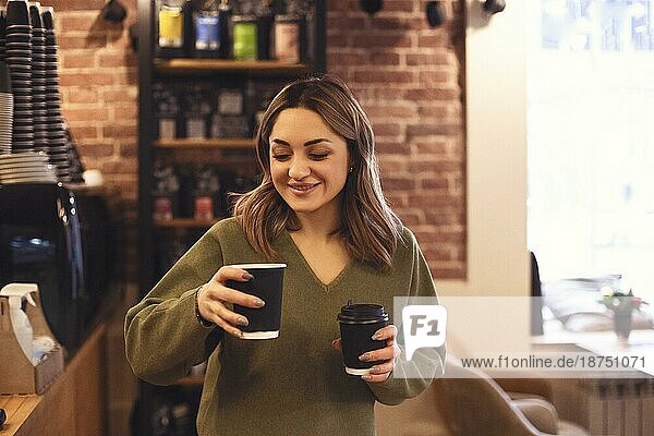 Innenaufnahme einer jungen lächelnden Frau  die zwei Tassen Kaffee zum Mitnehmen hält  während sie in einem Café steht  selektiver Fokus auf eine Frau  die heiße Getränke während eines morgendlichen Treffens mit einem Freund bringt