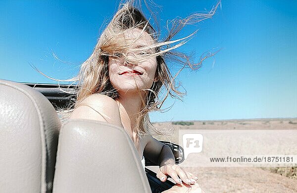 Fröhliche junge Frau in Sommerkleidung  die auf dem Beifahrersitz eines Cabrios sitzt und während einer Autofahrt über die Schulter in die Kamera schaut