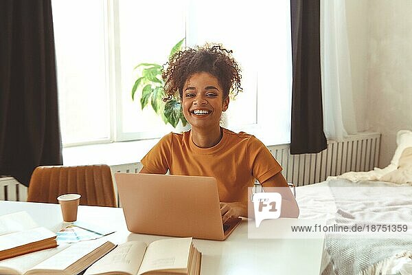 Fröhliche junge afroamerikanische Studentin  die zu Hause am Schreibtisch sitzt  umgeben von Lehrbüchern  vor dem Laptop fleißig lernt und sich online auf Prüfungen vorbereitet  mit einem zufriedenen Lächeln in die Kamera schaut