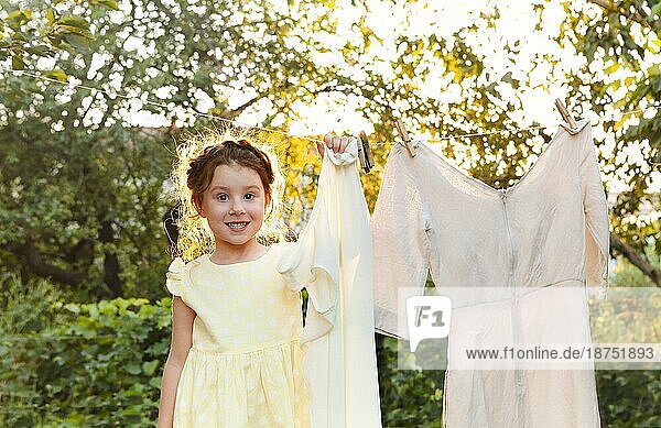 Niedliches kleines Mädchen im Kleid  das weiße Wäsche in einem Metallbecken im Garten wäscht  Kind  das bei der Hausarbeit hilft  Wäsche an der Wäscheleine aufhängt und sie im Garten trocknen lässt  Wäsche im Freien im Wind trocknet