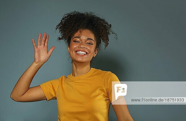 Schöne glückliche dunkelhäutige afrikanische amerikanische ethnische Frau im gelben TShirt die die Hand zur Begrüßung hebt  während sie mit einem angenehmen Lächeln in die Kamera blickt  posiert vor einem grauen  grünen Studiohintergrund